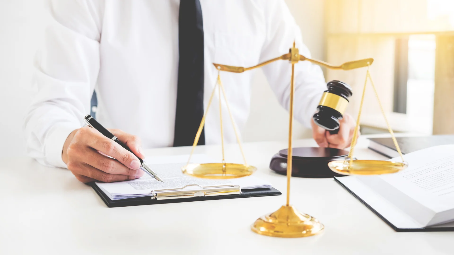 takım elbieli kişi bir elinde tokmak diğer elinde kalem ve önündeki evrağı iinceliyor.Evrağın önünde ise hukuk terazisi var