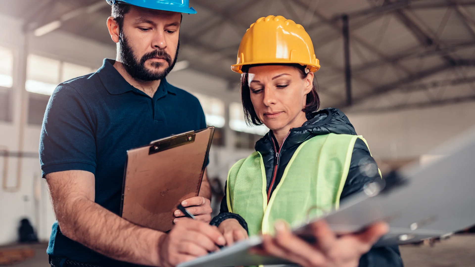 Erkeğin kafasında mavi baret elinde bir evrak ve kalem var.Kadının kafasında sarı baret ve üzerinde yelek elindeki evrağı erkeğeuzatıyor.